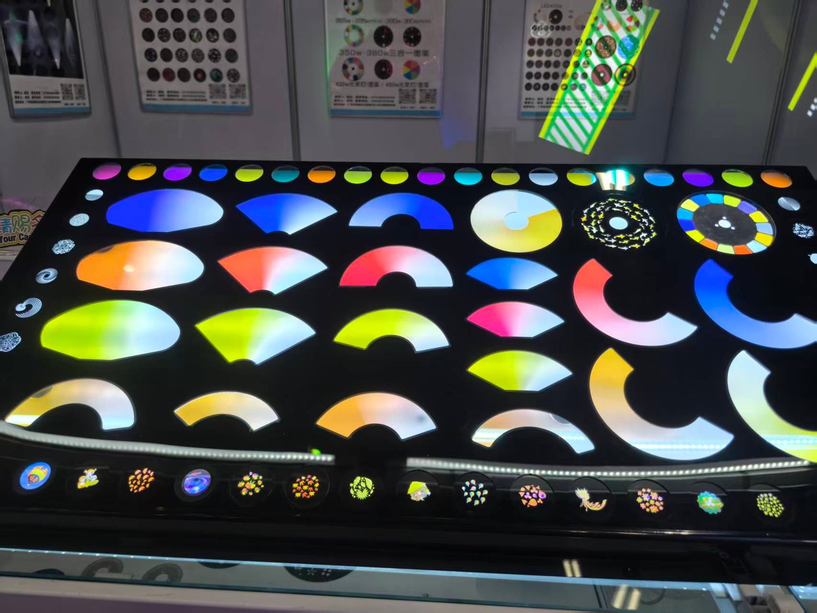 A display table features various colorful, illuminated semi-circular and circular patterns reminiscent of a stage light color wheel, with more pattern samples shown on vertical boards in the background.