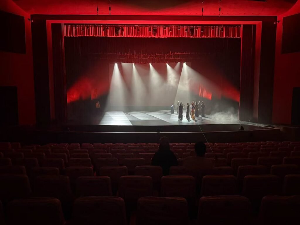 A dimly lit theater stage with dramatic red stage lighting, spotlights shining on a group of people standing together, and empty seats visible in the foreground.