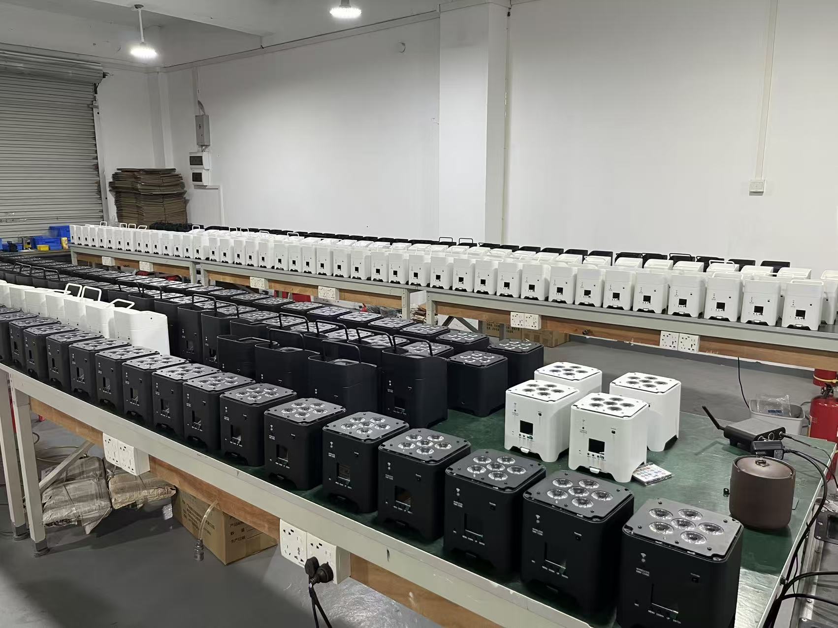How Many Battery Powered Uplights Do You Actually Need? 1 Rows of black and white electronic devices, including battery operated uplights, are neatly arranged on a long workbench in a factory or workshop setting with white walls and a concrete floor.